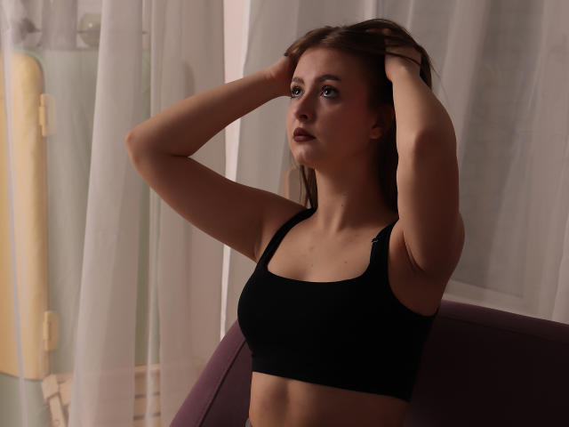 AdelPearson with hands in brown hair, wearing a black sports bra, showing her tits, has a serious expression, standing in front of a curtain, looking to the side.