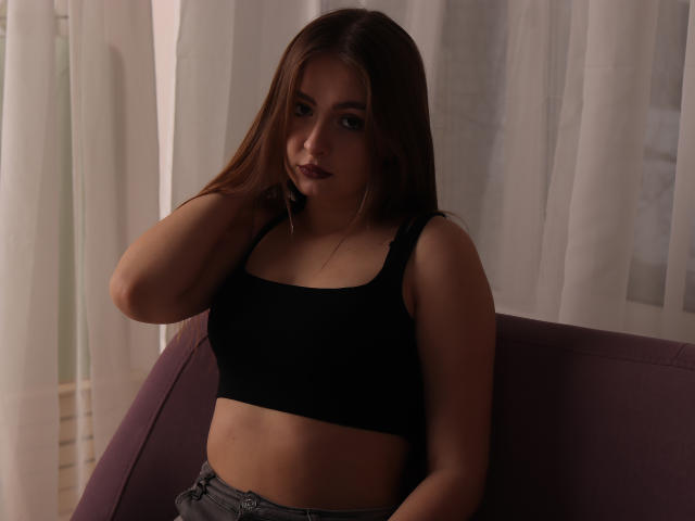 AdelPearson with long brown hair, wearing a black sports bra and grey pants, sitting on a purple chair, hand in hair, looking at camera with serious expression.