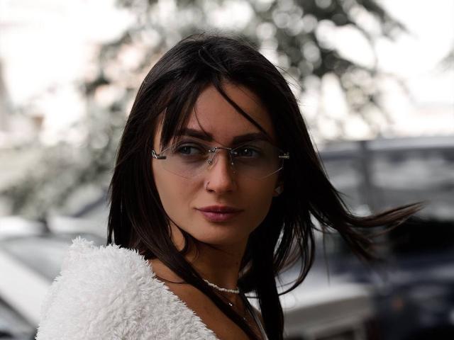 LikaLuis with dark brown hair, wearing clear glasses, white fluffy top, and pink lipstick, looking straight at camera, wind blowing slightly. She's got a cute, sexy face.
