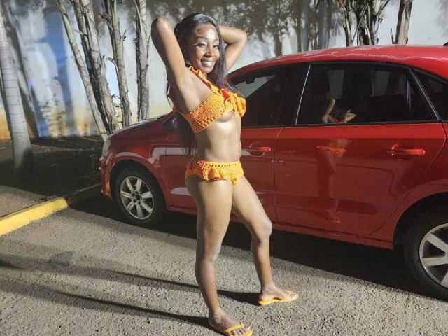 SkylerSeekret in an orange, ruffled bikini with her hands behind her head, standing in front of a red car, wearing yellow flip-flops, smiling. She's a dark-skinned woman.
