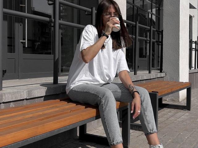 LikaLuis with brown hair, glasses, white shirt, gray jeans, drinking from a cup, sitting on a wooden bench, tattoo on right wrist, black wristband, gray pavement, daytime.