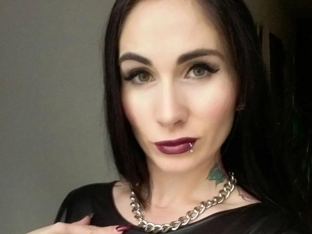 LadyPina-hot with dark brown hair, green eyes, black eyeliner, purple lipstick, pierced lower lip, silver chain necklace, black top, and visible tattoo on her right neck.