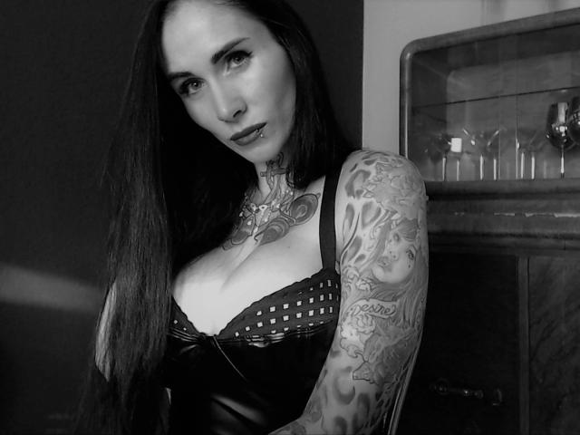 Black-and-white photo of LadyPina-hot with long dark hair, tattoos on her neck and arm, and large tits in a leather dress, looking seductively at the camera.