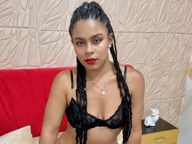 AlizonHoot with long braids, red lipstick, wearing black lace bra, necklace, sitting on red couch, tan textured wall, small white statue on yellow cabinet. Boobs, black bra, lace.