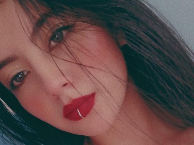 MonaSwan has red lipstick, slightly parted pinkish lips with a silver piercing, and long brown hair with strands in her face. She has blue eyes and a soft, fair skin.