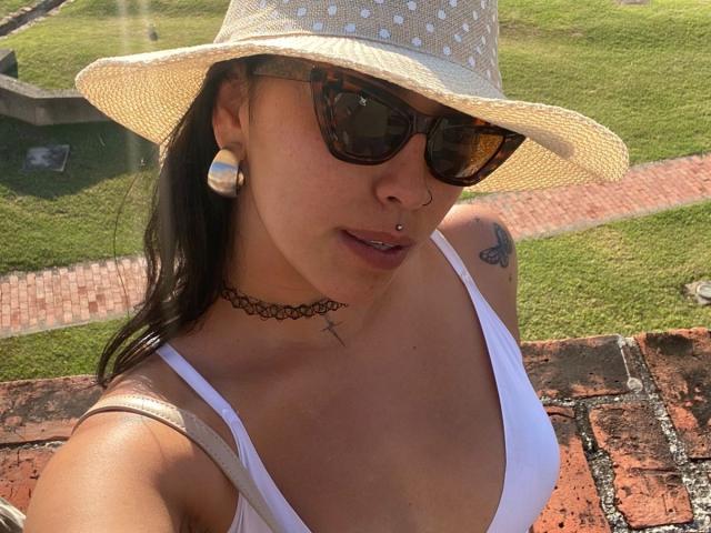 Elizabeth Macy, usando óculos de sol escuros, uma regata branca que mostra seus seios, um chapéu de palha, alargadores de orelha, um piercing no nariz, um colar e uma tatuagem, além de um piercing nos lábios, em pé na grama iluminada pelo sol..