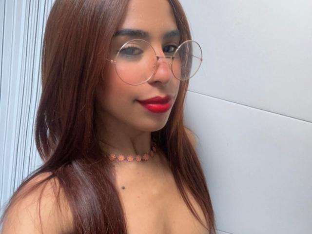 LisshaCute has brown hair, red lipstick, round glasses, and a flower necklace, showing off her small tits and cute face with a slight smile.