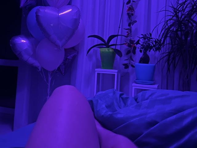 Trima's legs are in the foreground, with blue-purple lighting. Balloons and potted plants are in the background. Trima's bed has messy blue-gray sheets. Trima's pussy is not visible.