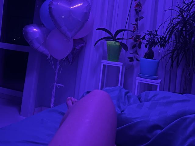 Trima's legs on a bed, blue sheets, purple light, white window, balloons, potted plants, and a plant hanging in the background. Trima's ass is partially visible.