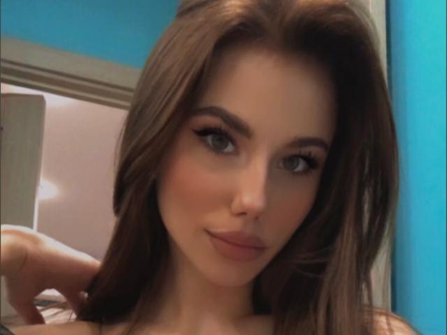 NicoleNight has long brown hair, green eyes, and a subtle smile. She's looking at the camera with a slight smirk, her hand behind her head. She's cute.
