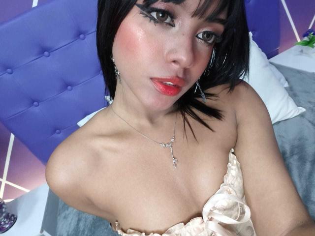 HannaCrow with short black hair, red lipstick, and eye makeup, wearing a strapless cream dress, showing her small tits, necklace, and earrings, on a bed with a blue headboard.