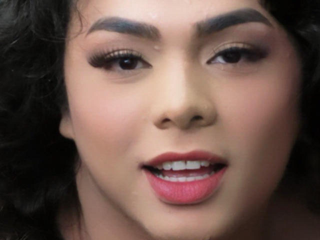 LiliithBuckland's face with full lips, pink lipstick, dark curly hair, thick eyebrows, and brown eyes. She's smiling slightly, showing her white teeth. Her skin is smooth and light.