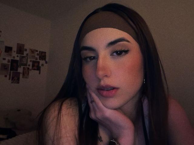 MiaHarperr with dark brown hair, headband, and eyeliner, resting her chin on her hand, looking seductively at the camera, her lips slightly parted. Dimly lit, her skin is flushed.