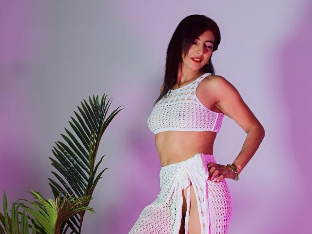 SamanthaSmithson with medium tits, wearing a white crocheted crop top and skirt, standing sideways, smiling, brown hair, palm leaf on left, pink-purple light.