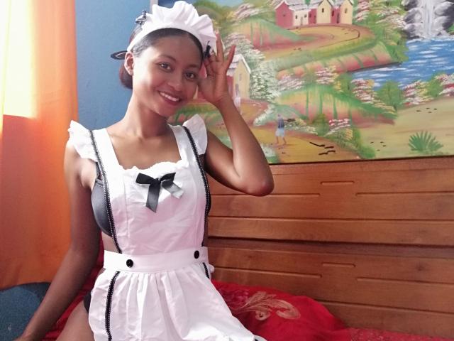 Cayrah, small dark-skinned woman with cute smile, wearing white maid outfit with black ribbon, raising right hand, standing in sunlit room, colorful painting in background.