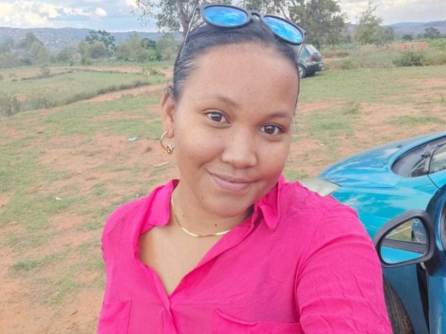 EmillyHottie with dark skin, small nose, and subtle smile, wearing a pink shirt, gold necklace, and blue sunglasses on her head, standing next to a blue car.