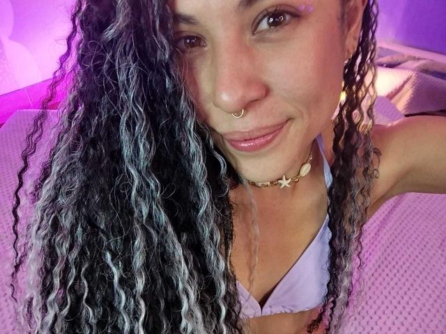 HellenK with long curly black and white hair, nose ring, star choker, pink lips, smiling, wearing a lilac bikini top, purple lighting, bed in background.