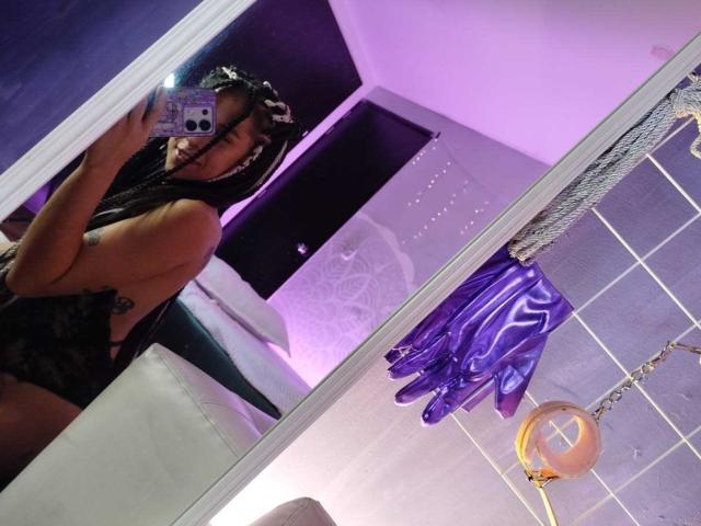 HellenK with long braids, wearing black lace lingerie, taking a mirror selfie, showing her ass and purple gloves, in a room with purple lighting.