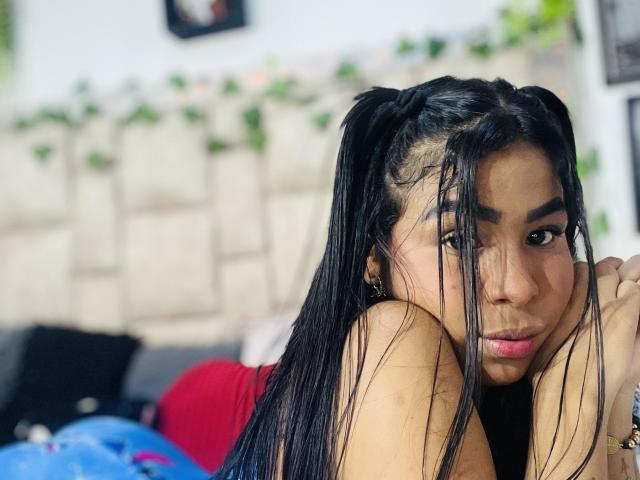 MollyXDamiam has long black hair in pigtails, looking over her shoulder with pouty lips, dark eyebrows, and a slight nose, showing her bare shoulders and a red top.