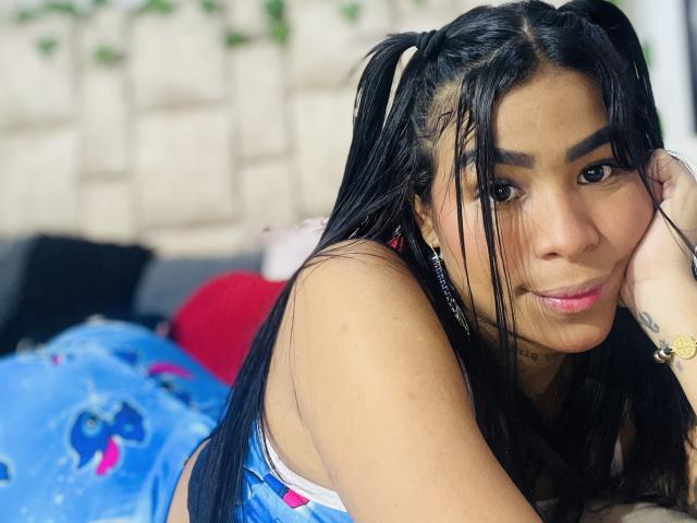 MollyXDamiam with long black pigtails, brown eyes, and tan skin, lying on a bed in a blue dress, smiling subtly, showing a tattoo on her right wrist.