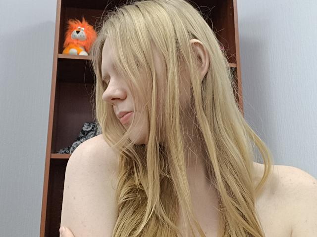 Blonde Laliina with long hair, smiling softly, topless, her small tits barely visible, standing in front of a wooden shelf with a fluffy orange toy.