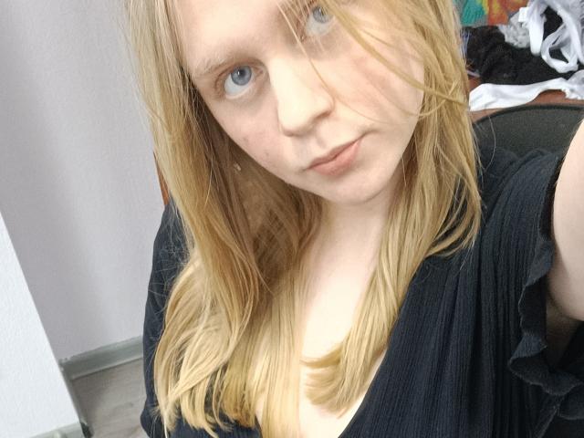 Laliina has blonde hair, blue eyes, and fair skin. She's wearing a black top with ruffled shoulders, looking into the camera with a slight, confident smirk.