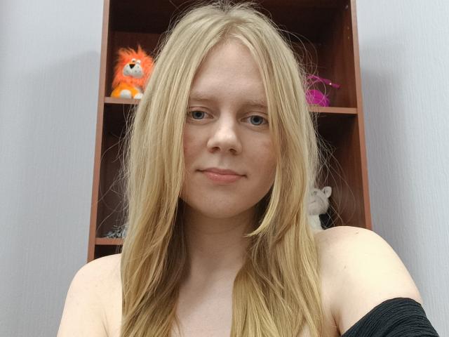 Blond Laliina with fair skin and blue eyes, bare shoulders, black strap, slight smile, and a cute orange plushie in the background.