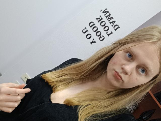 Laliina with blonde hair, blue eyes, and a serious face, wearing a black top with a low neckline, showing her small boobs, in a simple room.