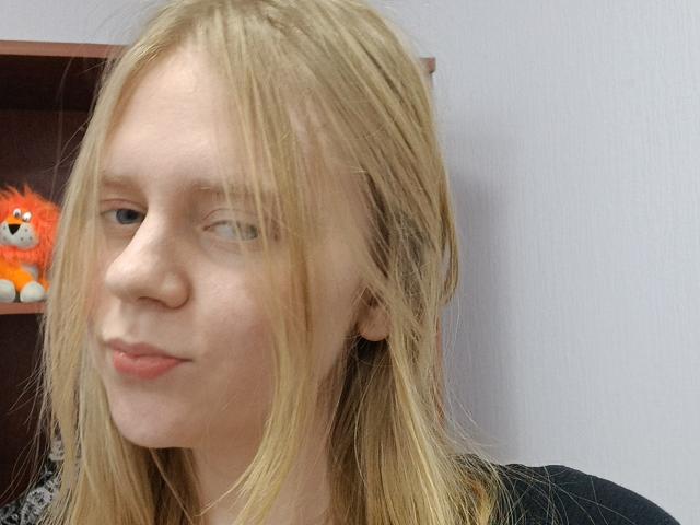 Laliina has blonde, slightly messy hair, pale skin, and light blue eyes. She's looking to the side with a subtle, closed-lip smile. Her small, pinkish lips are slightly parted.