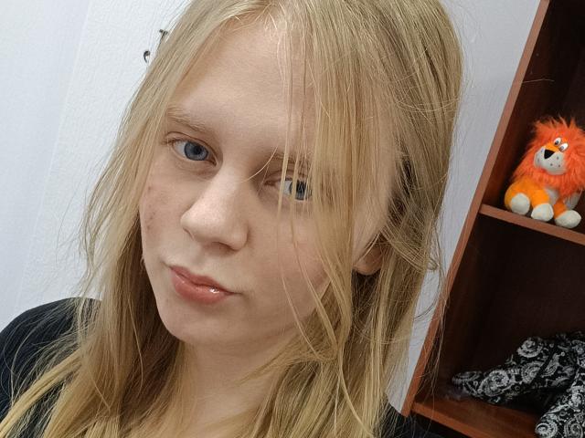 Laliina has blonde hair, blue eyes, and a small lip piercing. She's wearing a black top, with a red and orange stuffed animal in a wooden shelf behind her.