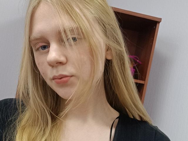 Laliina has long blonde hair, fair skin, and blue eyes. She's wearing a black top with a visible black strap, looking slightly bored with a subtle pout.