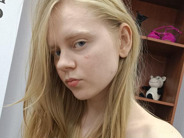 Blond Laliina with pale skin, small zit on right cheek, and pouty lips, wearing nothing, showing bare shoulders, standing near a wooden shelf with a stuffed monkey.
