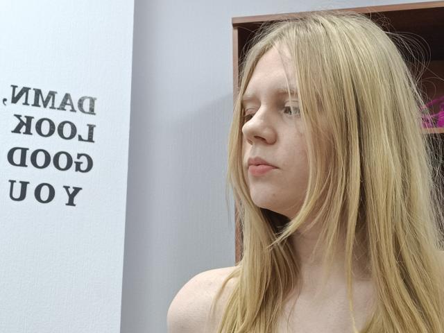Laliina with long blonde hair, pale skin, and pouty lips, looking to the side, topless, showing her bare shoulders. She has a serious, slightly annoyed expression.