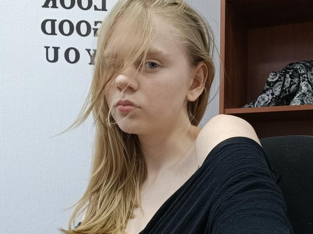 Laliina has blonde hair covering her right eye, a serious expression, and a black off-shoulder top, showing her bare shoulder and upper chest. She's in a chair.
