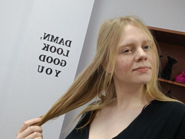 Laliina has blonde hair, fair skin, small tits, and a slight smile, wearing a black top, holding her hair with one hand, looking to the side.