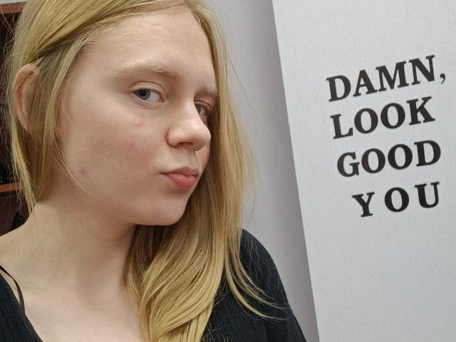 Laliina has pale skin, blonde hair, and a pouty face with small zits on her nose. She's wearing a black shirt, looking slightly annoyed.