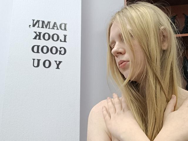 Laliina has long blonde hair, light skin, and is topless, covering her small tits with her hands, looking to the side with a subtle smile.