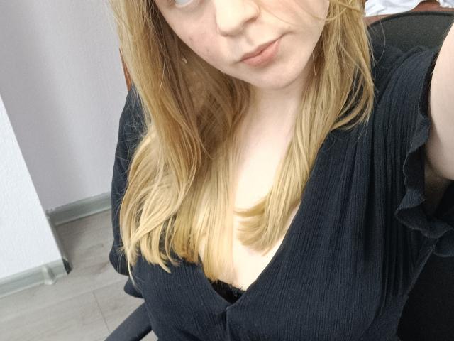 Laliina with blonde hair, wearing a black ribbed dress, showing her large tits in a black bra, slightly tilted head, and a subtle smile, in a selfie.