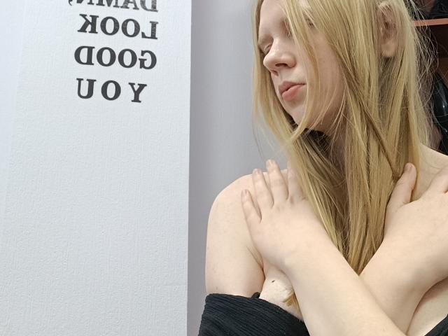 Laliina with blonde hair, eyes closed, black off-shoulder top, hands touching her chest, looking soft and calm. Boobs visible, subtle and natural. She's slightly turned, looking right.
