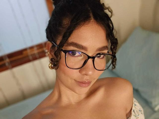 KendallPiierce69 has curly black hair, wearing black glasses, with a cute smile, and is topless, showing her bare shoulders and a small gold earring in her right ear.