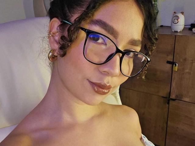 KendallPiierce69 with curly brown hair, glasses, shiny brown lips, gold hoop earrings, and off-shoulder top, showing bare shoulders, sitting in a dimly lit room.
