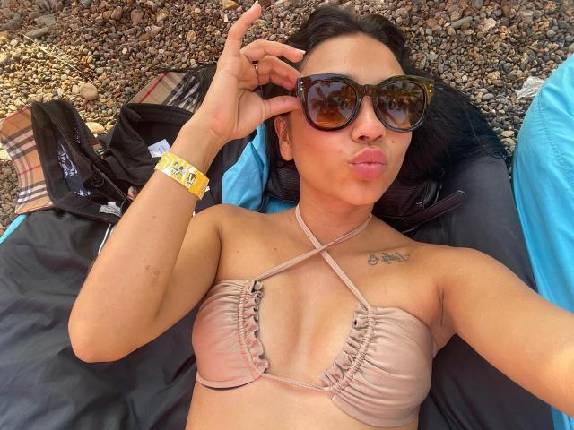 Sophie Denisee, acostada en una playa, vistiendo un top de bikini color piel con bordes fruncidos, gafas de sol negras y una pulsera amarilla, haciendo una mueca, con tatuajes en el pecho..