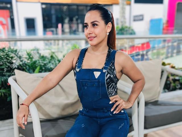 BombSophie with brown hair in a ponytail, wearing blue denim overalls, sitting on a bench, smiling, hand on hip, showing small tits, cute, tan skin, earrings.