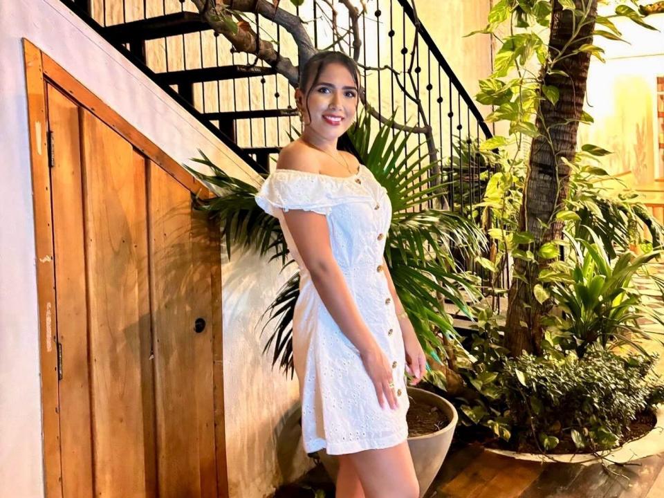 SofiaAlejandraX stands in a white off-shoulder dress with buttons, smiling, near a wooden door and black iron staircase. She has dark hair, green eyes, and small tits.