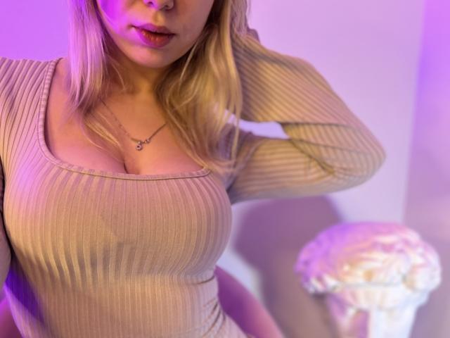 ElsaStormy with blonde hair, wearing a ribbed, beige, long-sleeve top, showing her big tits, and a small necklace, under purple and blue lighting. A white statue is in the background.