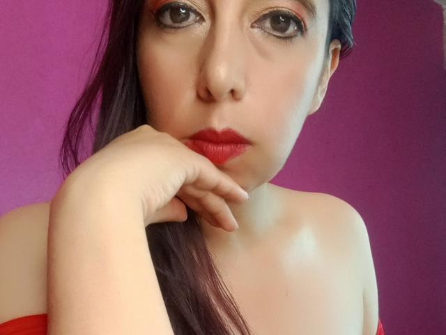 SublimeMature with red lipstick, dark eye makeup, and hand on chin, has pale skin and long brown hair, wearing an off-shoulder red top, looking directly at the camera.