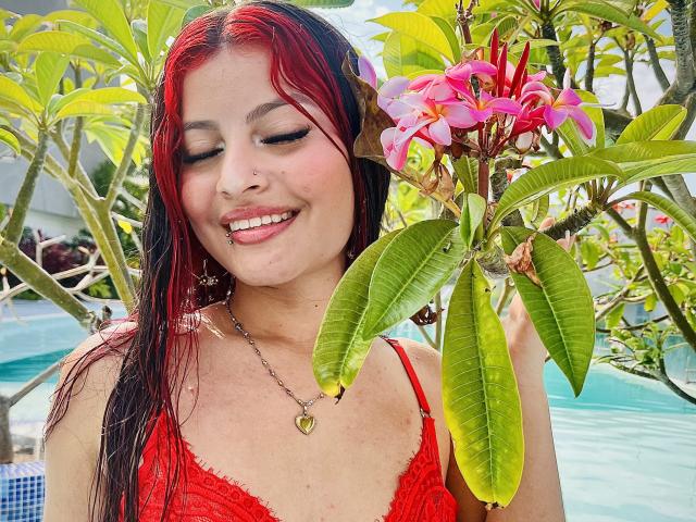 AshleyFostter69 with red and black wet hair, smiling, holding pink flowers, wearing red lace top, heart necklace, and nose ring, standing by a blue-tiled pool.
