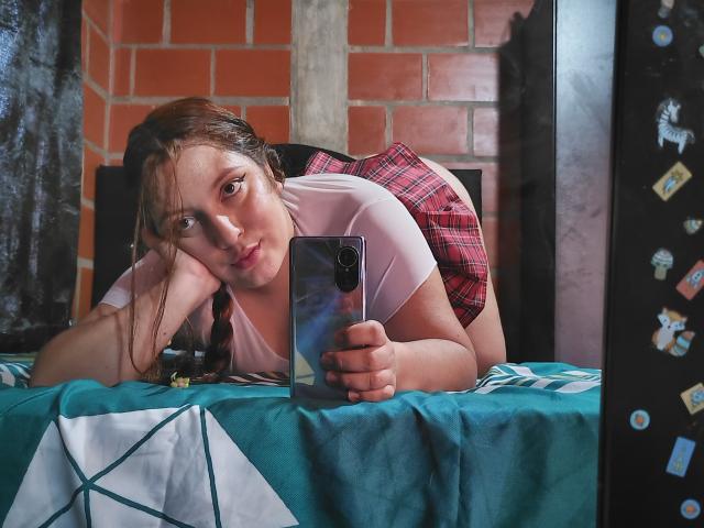 CaroFoxy, avec des cheveux bruns, un t-shirt blanc, une jupe à carreaux rouges, allongée sur un lit, prenant un selfie avec son téléphone, regardant l'appareil photo, une housse de lit à motifs verts, reflet dans un miroir, expression légèrement boudeuse..