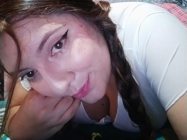 CaroFoxy, avec ses tresses brunes, son haut blanc et son eye-liner ailé, appuyée sur sa main, souriant avec ses lèvres roses, son menton reposant sur son poignet, montrant légèrement ses seins..