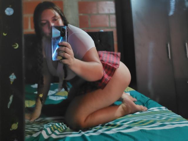 CaroFoxy, avec des cheveux tressés, un haut blanc, une jupe à carreaux rouges, et le cul nu, est agenouillée sur le lit, prenant un selfie devant un miroir avec son téléphone, montrant ses fesses et son dos dans le reflet du miroir..